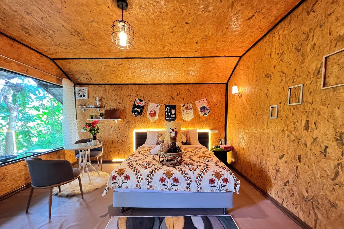 A bedroom with a bed, a desk, and a chair. There is a window with a view of trees.