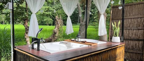 A wooden outdoor hot tub with a white basin and black faucets, surrounded by a bamboo fence and lush greenery.