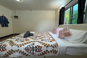 Un dormitorio con cama, almohadas, una manta, una ventana con cortinas y un aire acondicionado instalado en la pared.
