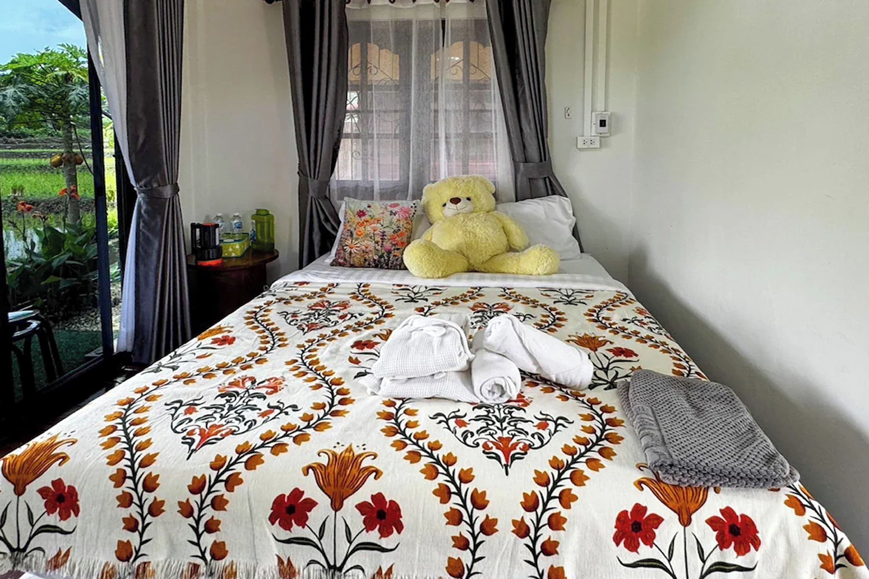 Un dormitorio con cama, un oso de peluche grande, una ventana con cortinas y vistas a la vegetación.