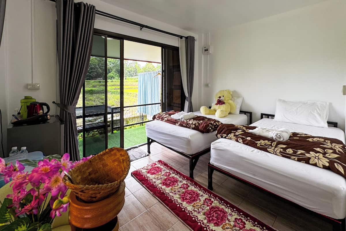 A hotel room with two beds, a large window with curtains, a teddy bear on one bed, and a small table with a kettle and cups.