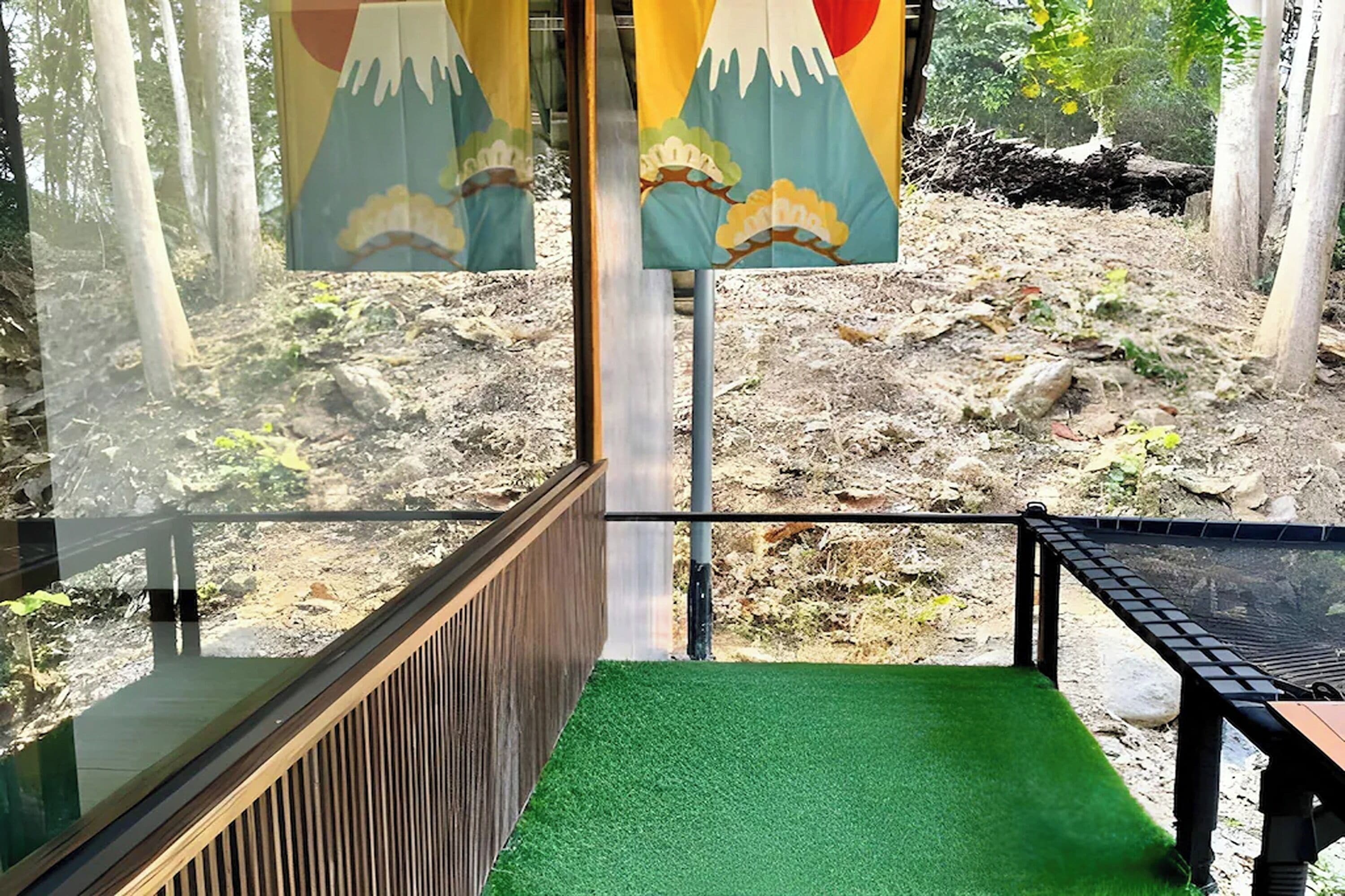 Vista a través de un recinto de vidrio con barandilla de madera, suelo de césped artificial verde y zona exterior rocosa con árboles.