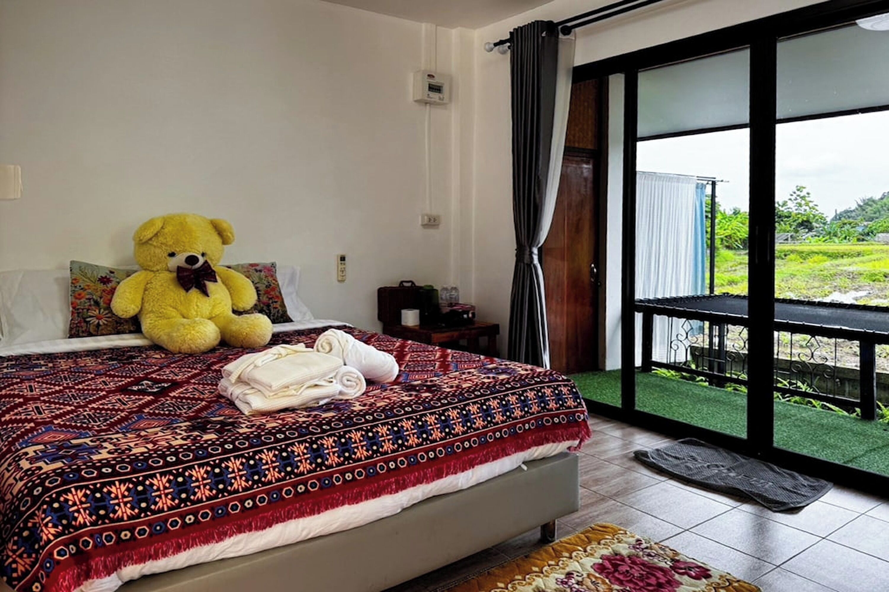 A hotel room with a large bed, a teddy bear on the bed, a view of a green landscape through the window, and a balcony with a railing.
