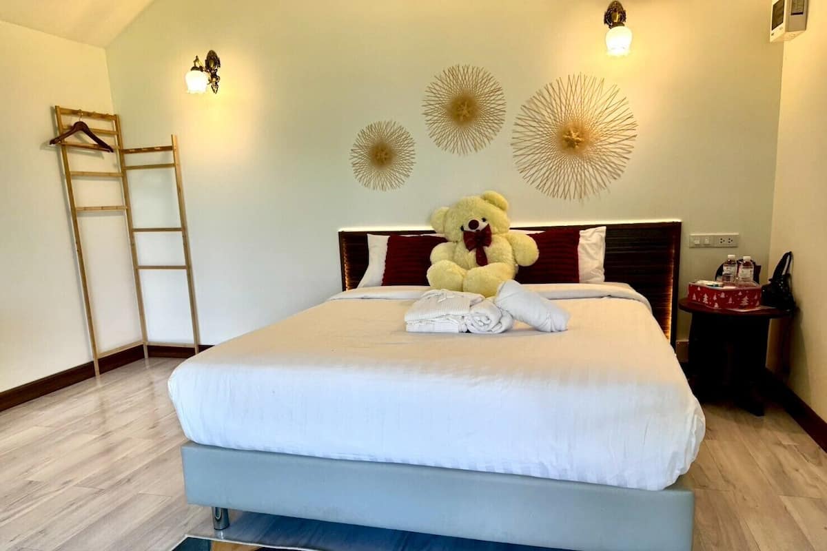 A neatly made bed with a teddy bear and folded towels, a wooden ladder, decorative wall sconces, and a small table with a bottle and a box.