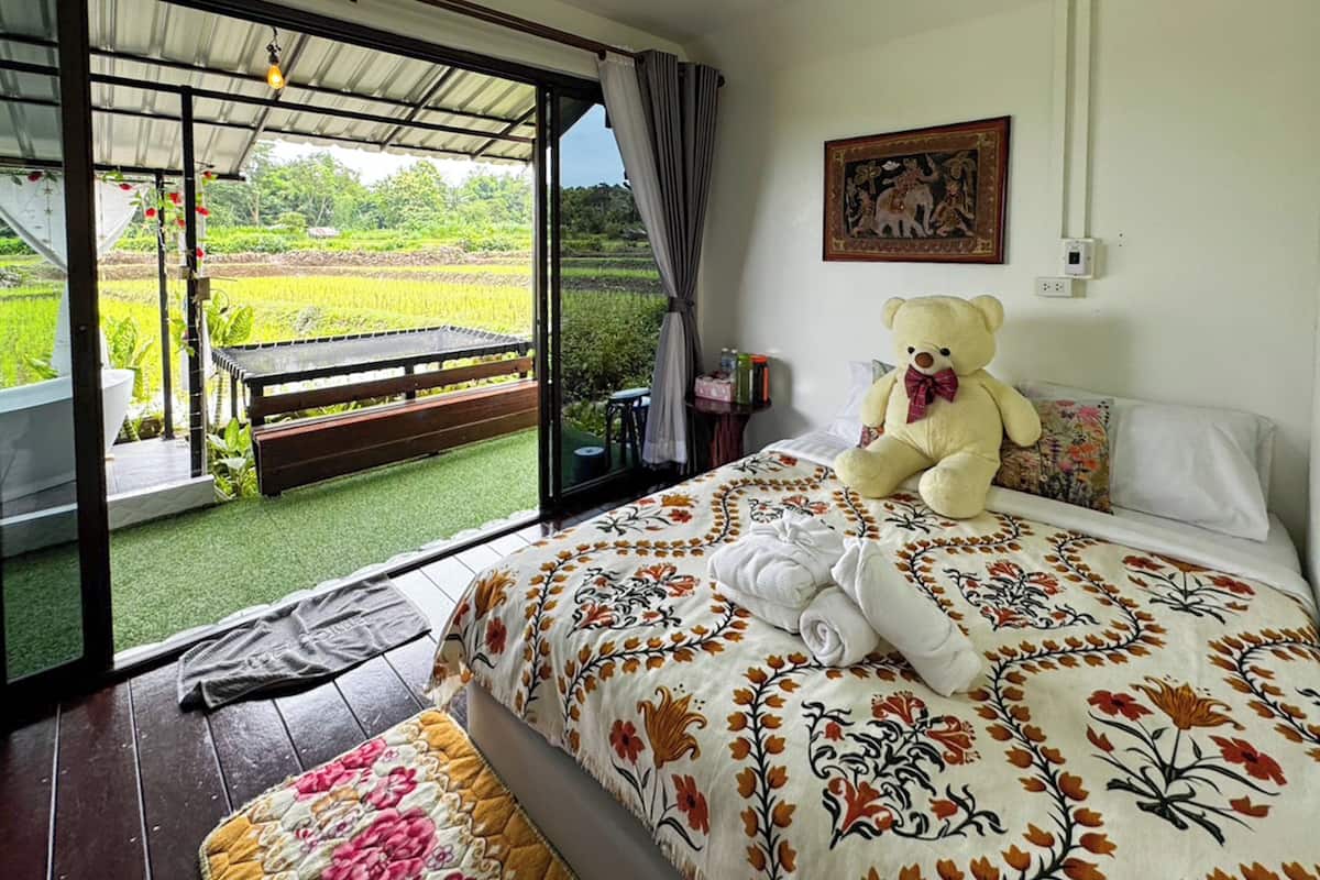 A bedroom with a bed, a large teddy bear, a painting, and a view of a green landscape through an open door.