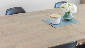 Una mesa de madera con un recipiente blanco y un florero de flores blancas sobre un mantel individual azul.