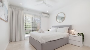 Une chambre moderne avec un lit gris, des tables de chevet blanches, un ventilateur de plafond et une grande fenêtre avec des stores.