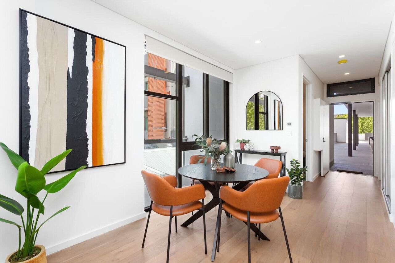 Ein moderner Essbereich mit einem runden Tisch und orangefarbenen Stühlen, einem großen abstrakten Kunstwerk an der Wand und Blick auf ein Backsteingebäude durch große Fenster.