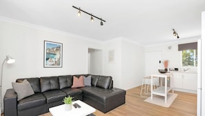 A modern living room with a black leather sectional sofa, a white coffee table, and a small side table. There is a kitchen area with white cabinets and a window.