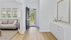 Un couloir avec un canapé de couleur claire, une console blanche agrémentée d’un miroir et une porte donnant sur un jardin.