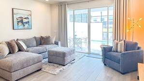 Ein modernes Wohnzimmer mit einem grauen Ecksofa, einem kleineren grauen Sessel und einer Glastür, die auf einen Balkon führt.