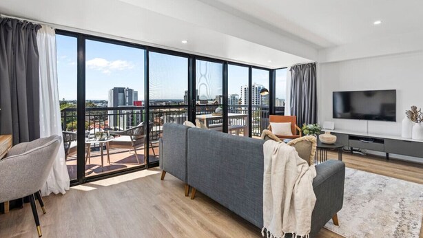 Un salon moderne avec une grande télévision, un canapé gris et un balcon offrant une vue sur la ville.