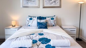 Un lit bien fait, recouvert d’une couvre-lit à motifs floraux, avec deux oreillers à rayures bleues et blanches, et une lampe de chevet posée sur une table de nuit.