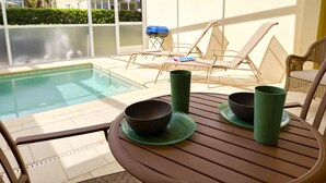 Un espace aménagé au bord de la piscine, avec des chaises longues, une table sur laquelle sont disposés des bols et des tasses, et une vue sur une maison à travers une paroi vitrée.