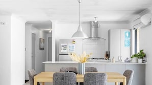 A modern kitchen with white cabinets, a central island, and a dining area with a wooden table and grey upholstered chairs. - Jindee Beach 2km away | Magical memories in Butler (Butler)