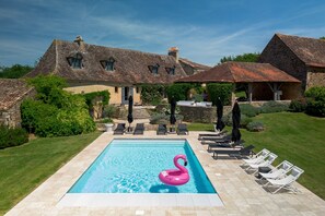 Une piscine avec un flotteur en forme de flamant rose, des chaises longues et un bâtiment en pierre surmonté d’un toit en tuiles.