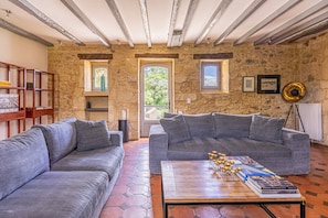 Una habitación con paredes de piedra, techo de madera, un sofá gris, una mesa de centro con libros y una ventana con vistas.