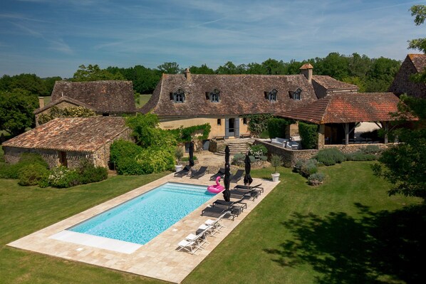 Une grande piscine entourée de chaises longues et de parasols, elle-même bordée de bâtiments en pierre aux toits de chaume.