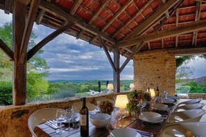 Un espace extérieur aménagé pour les repas, avec un mur en pierre, des poutres en bois et une vue sur la campagne.