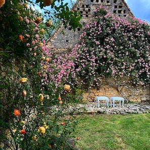 Ein Steinhaus mit einem Garten davor, der rosa und gelbe Blumen sowie zwei weiße Stühle zeigt.