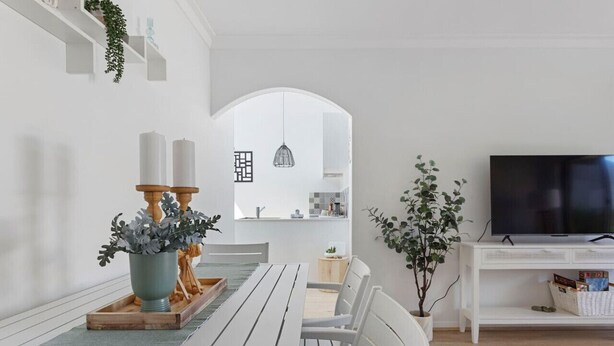 A modern living room with a wooden dining table, white chairs, a flat-screen TV, and a kitchen area visible through an archway. - Bright & Stylish by the Sea! (Scarborough)