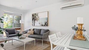 A modern living room with a grey sofa, white coffee table, and a dining table with white chairs. There is a large window with a view of greenery outside, and an air conditioning unit on the wall. - Bright & Stylish by the Sea! (Scarborough)