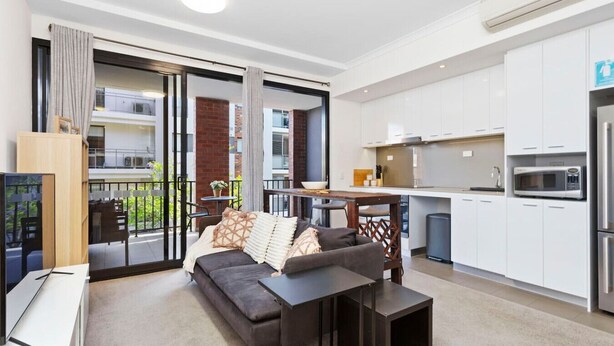 Un apartamento moderno con un sofá gris, una pequeña mesa de comedor, una cocina con armarios blancos y un balcón con vistas a la ciudad.