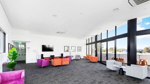 Un amplio salón con televisor de pantalla plana, varias opciones de asientos, incluyendo sillas anaranjadas y púrpuras, y grandes ventanales con vistas a la ciudad.