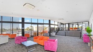 Un espacio interior amplio con ventanas grandes, varias zonas de estar con sillas de colores y una escalera que conduce a un nivel superior.