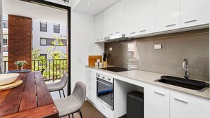 Una cocina moderna con armarios blancos, encimera negra y horno empotrado. Cuenta con un comedor con mesa y sillas de madera, y se aprecia un balcón con plantas y un edificio al fondo.