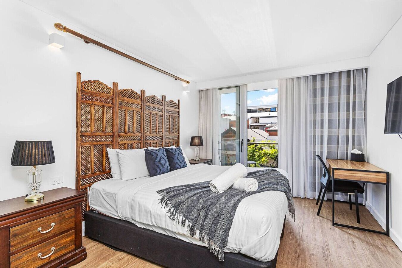 A modern hotel room with a large bed, a desk, and a balcony with a city view.