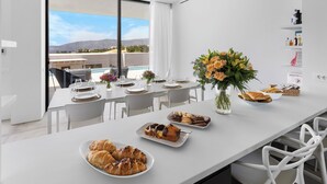 Un comedor moderno con vistas a una piscina y montañas, mobiliario blanco y una variedad de pasteles y flores sobre la mesa.
