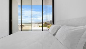 Un dormitorio con una cama grande, un ventanal corredero que da a un balcón con vistas al cielo y edificios lejanos.