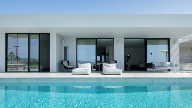 Una casa moderna con piscina, mobiliario blanco y amplios ventanales que dan a una sala de estar.