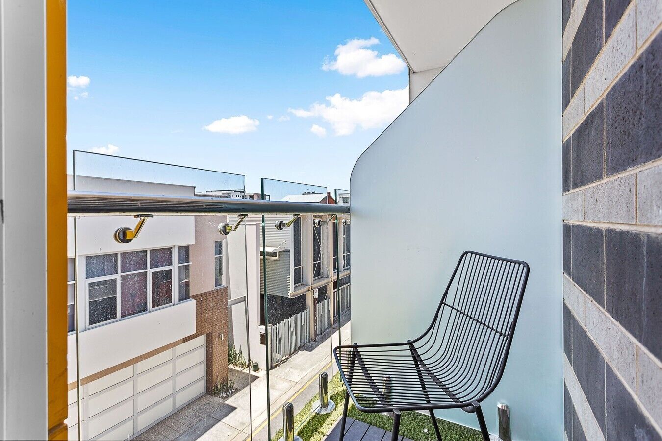 Un balcon étroit avec une chaise en métal noir, une porte vitrée et une vue sur les immeubles voisins.