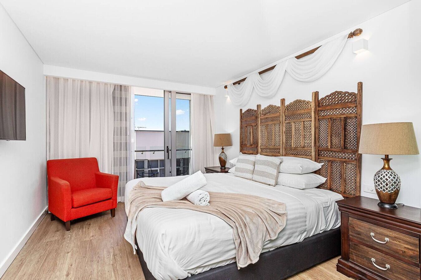 A bedroom with a large bed, a red chair, a wooden headboard, and a balcony with curtains.