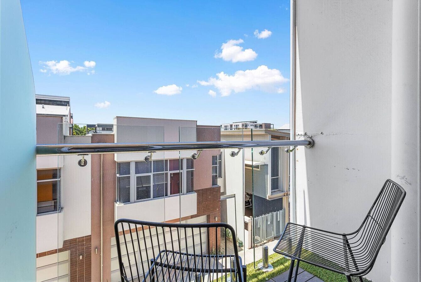 A balcony with a metal chair and a glass railing overlooking a residential area with modern buildings.