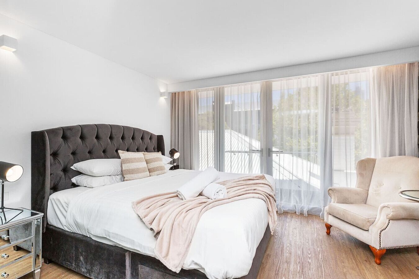 A modern bedroom with a tufted headboard bed, a bedside table with a lamp, a chair, and large windows with sheer curtains.