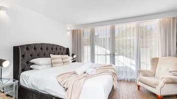 A modern bedroom with a tufted headboard bed, a bedside table with a lamp, a chair, and large windows with sheer curtains.