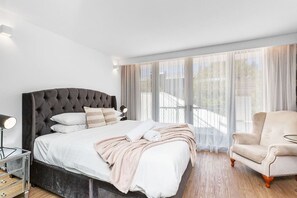 A modern bedroom with a tufted headboard bed, a bedside table with a lamp, a chair, and large windows with sheer curtains. - Nest on Newcastle – E7 (Perth)