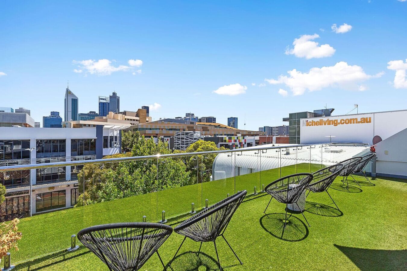 A rooftop terrace with artificial grass, black wireframe chairs, and a city skyline in the background.