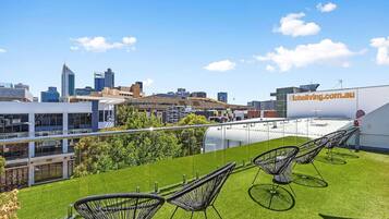 A rooftop terrace with artificial grass, black wireframe chairs, and a city skyline in the background.