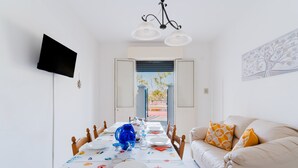 Een eetgedeelte met een lange tafel gedekt voor een maaltijd, een bank met kussens, een aan de muur bevestigde televisie en een deur die naar buiten leidt.