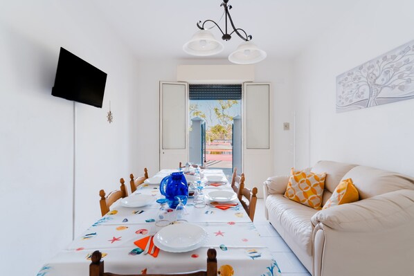 A dining area with a long table set for a meal, a sofa with cushions, a television mounted on the wall, and a door leading outside.