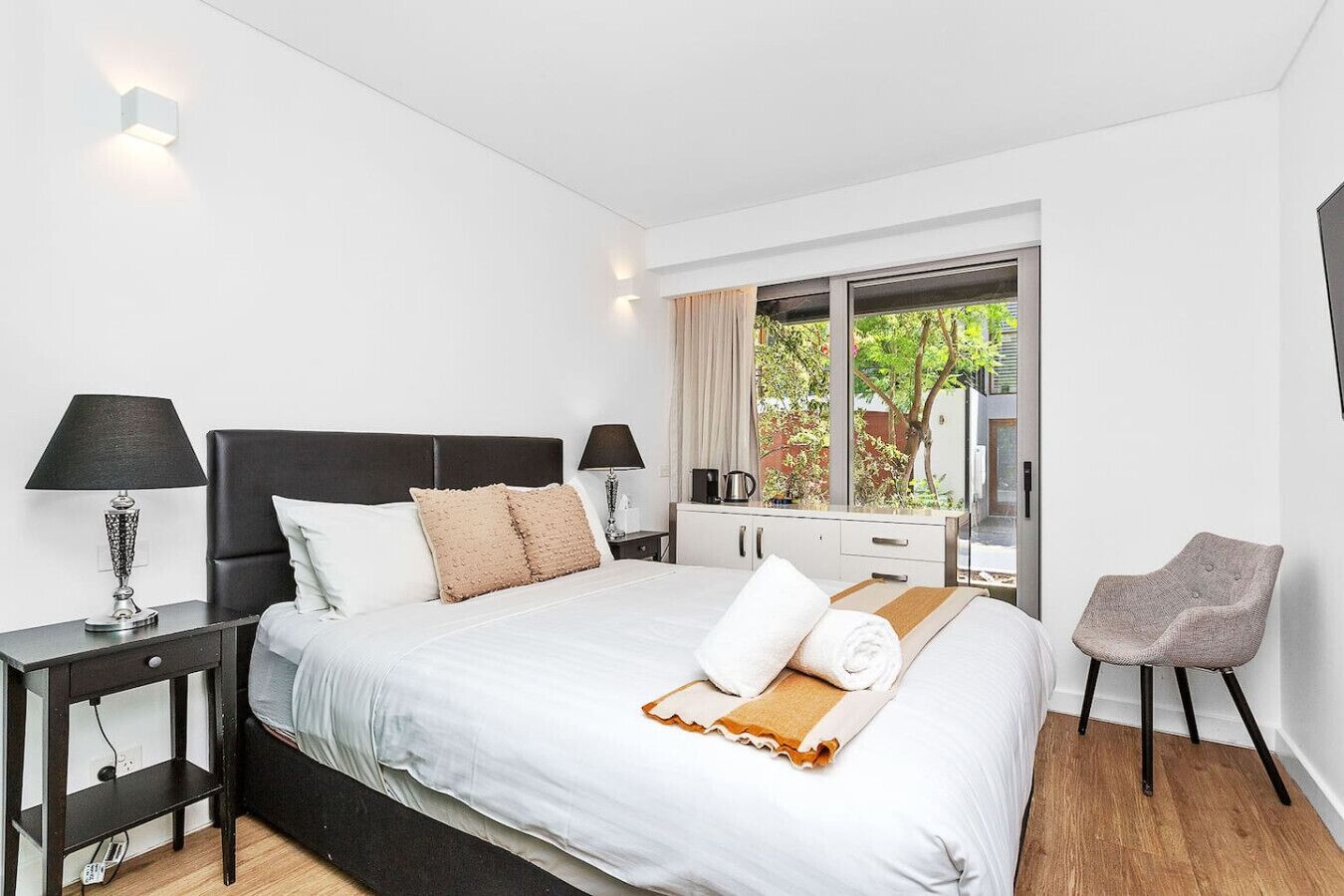 Een moderne hotelkamer met een groot bed, twee nachtkastjes met lampen, een dressoir, een stoel en uitzicht op groen via een schuifdeur van glas.