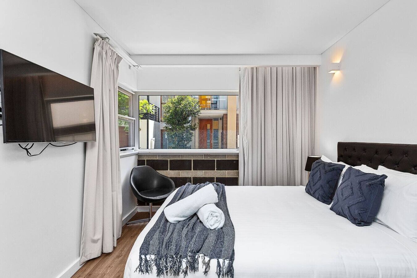 A hotel room with a bed, a TV mounted on the wall, a chair, and a window with curtains.