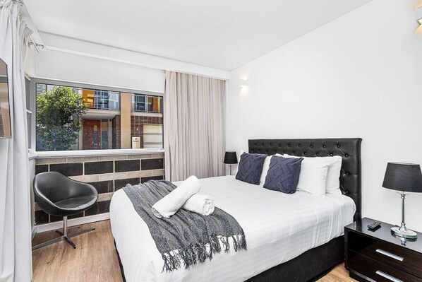 A modern hotel room with a large bed, a chair, a nightstand, and a window with a view of buildings.
