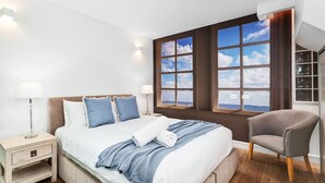 A modern bedroom with a large bed, two bedside tables, a chair, and a view of the sea through large windows. - Nest on Newcastle – T3 (Perth)