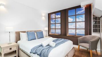 A modern bedroom with a large bed, two bedside tables, a chair, and a view of the sea through large windows.