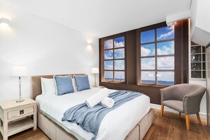 Un dormitorio moderno con una cama grande, dos mesitas de noche, una silla y vistas al mar a través de amplios ventanales.